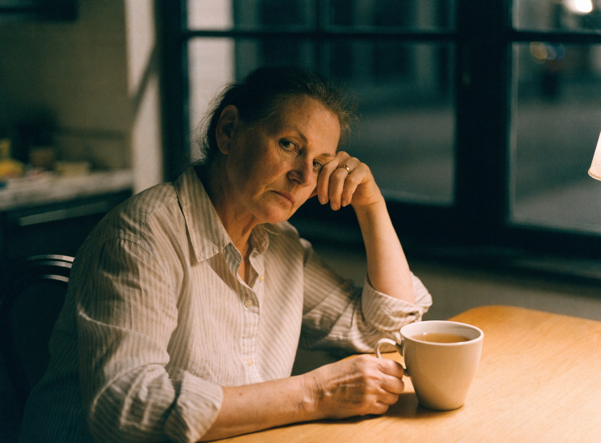 A woman in her fifties sitting alone at a kitchen table late at night with a cup of tea