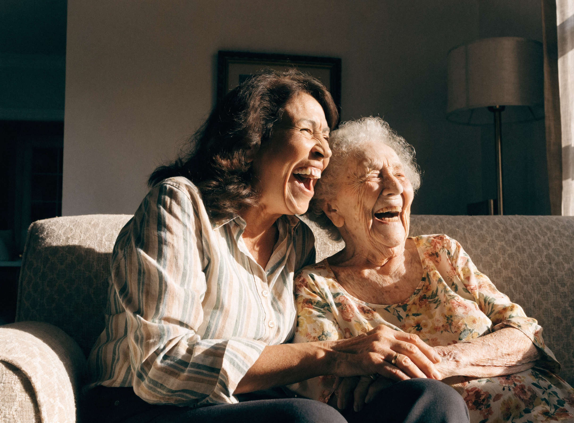 Adult daughter and her elderly mother laughing together on a living room sofa